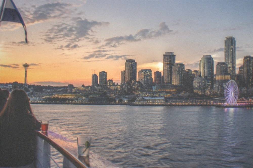 An open deck boat passing the Seattle skyline at dusk, with lit high-rise buildings and a ferris wheel along the waterfront