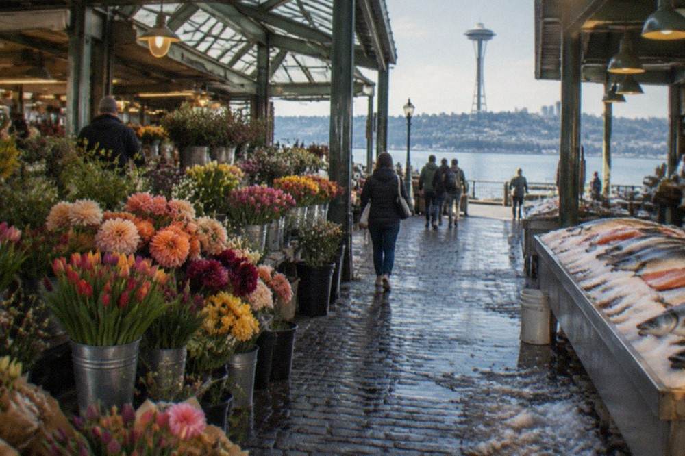 Flower stalls and produce stalls inside an open-air market with a bay visible beyond the roofline