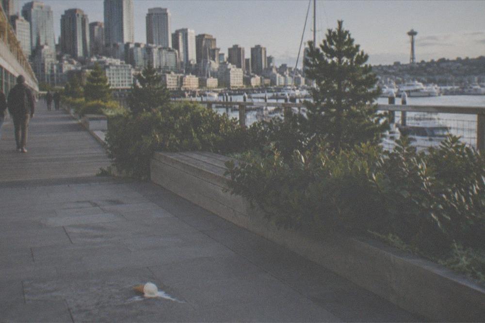 A waterfront promenade with park seating, public art, and moored boats along Elliott Bay.