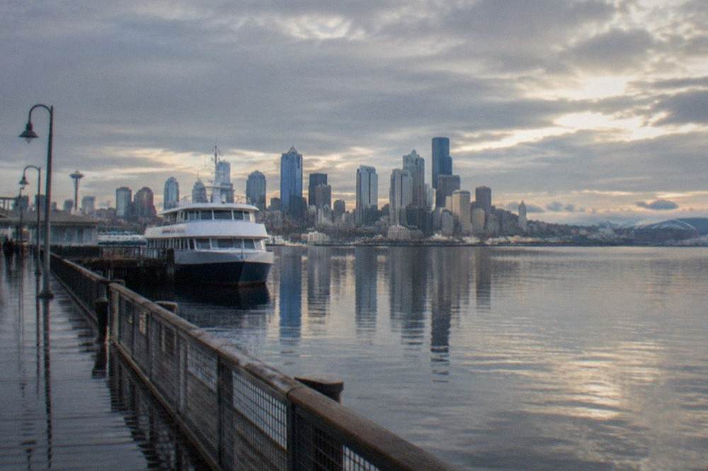 Seattle skyline across Elliott Bay under clearing skies after rain, with calm water in the foreground