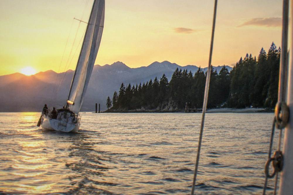 A sailboat heeling on Puget Sound under a bright late evening sky with the sun low over the water